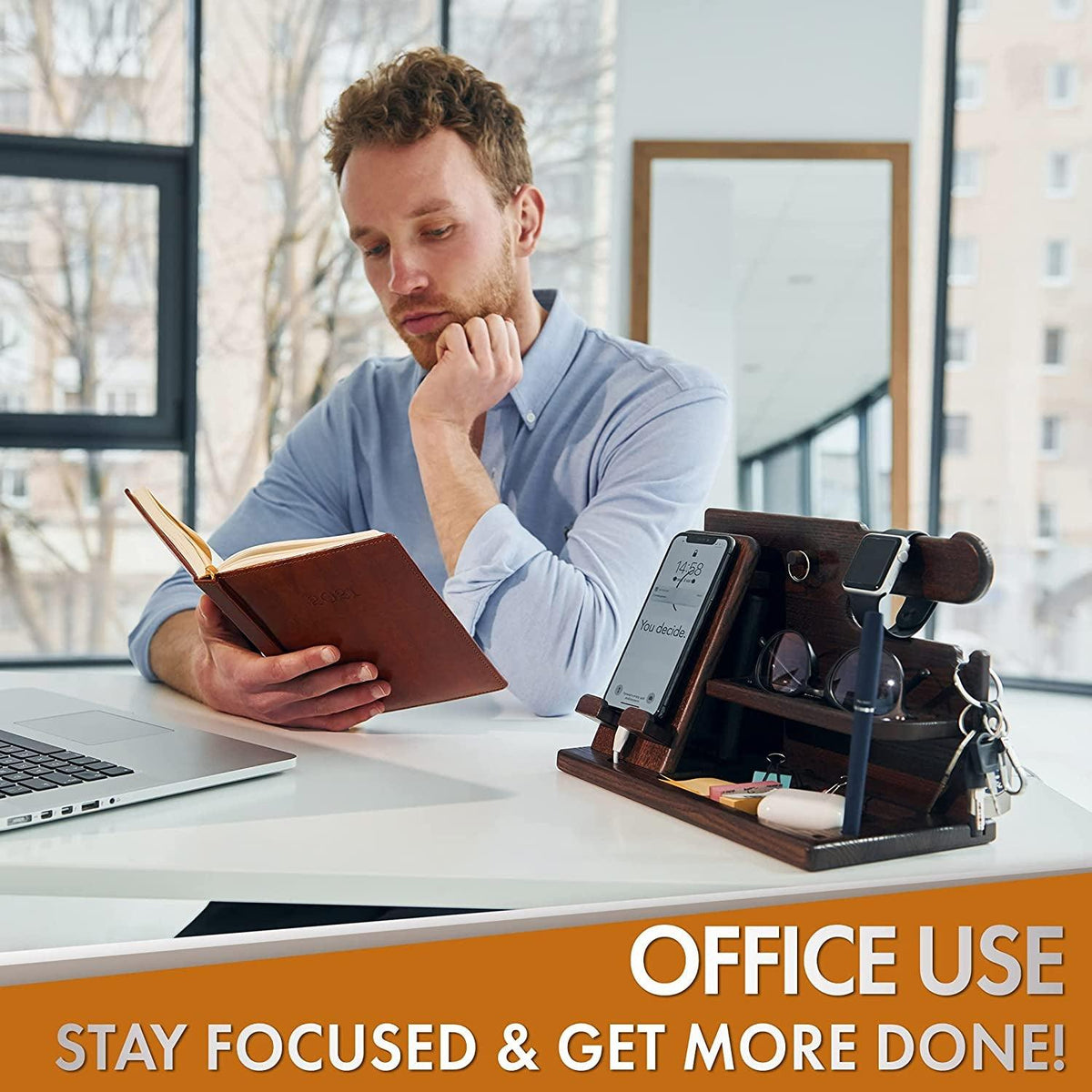 Man reading a book at a desk with a brown wood phone docking station and desk organizer.